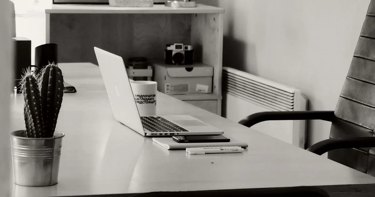 Bancada de escritório com notebook aberto, bloco de anotações e um vaso de metal com cacto, ao lado de cadeiras vazias.